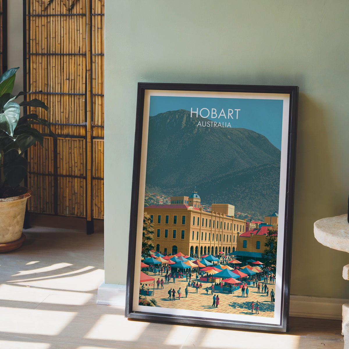 Tasmania city print in a black frame leaning on pale wood floor by a sage green wall and bamboo screen