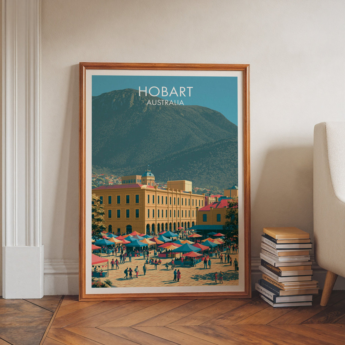Tasmanian travel artwork in a walnut frame on herringbone floor beside book stacks and a pale armchair