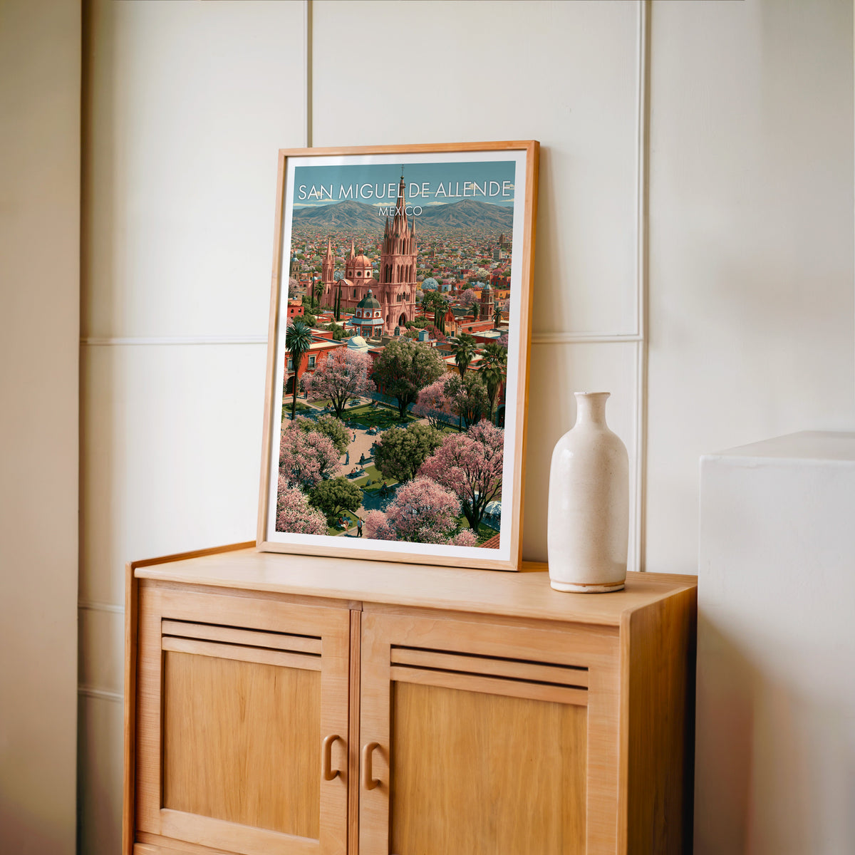 Mexican town artwork in an oak frame on a light oak cabinet against a cream panelled wall, tall vase