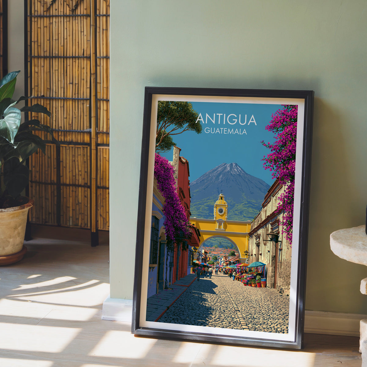 Cobbled street to the Santa Catalina arch, black frame leaning on pale wood floor by a sage wall and bamboo screen