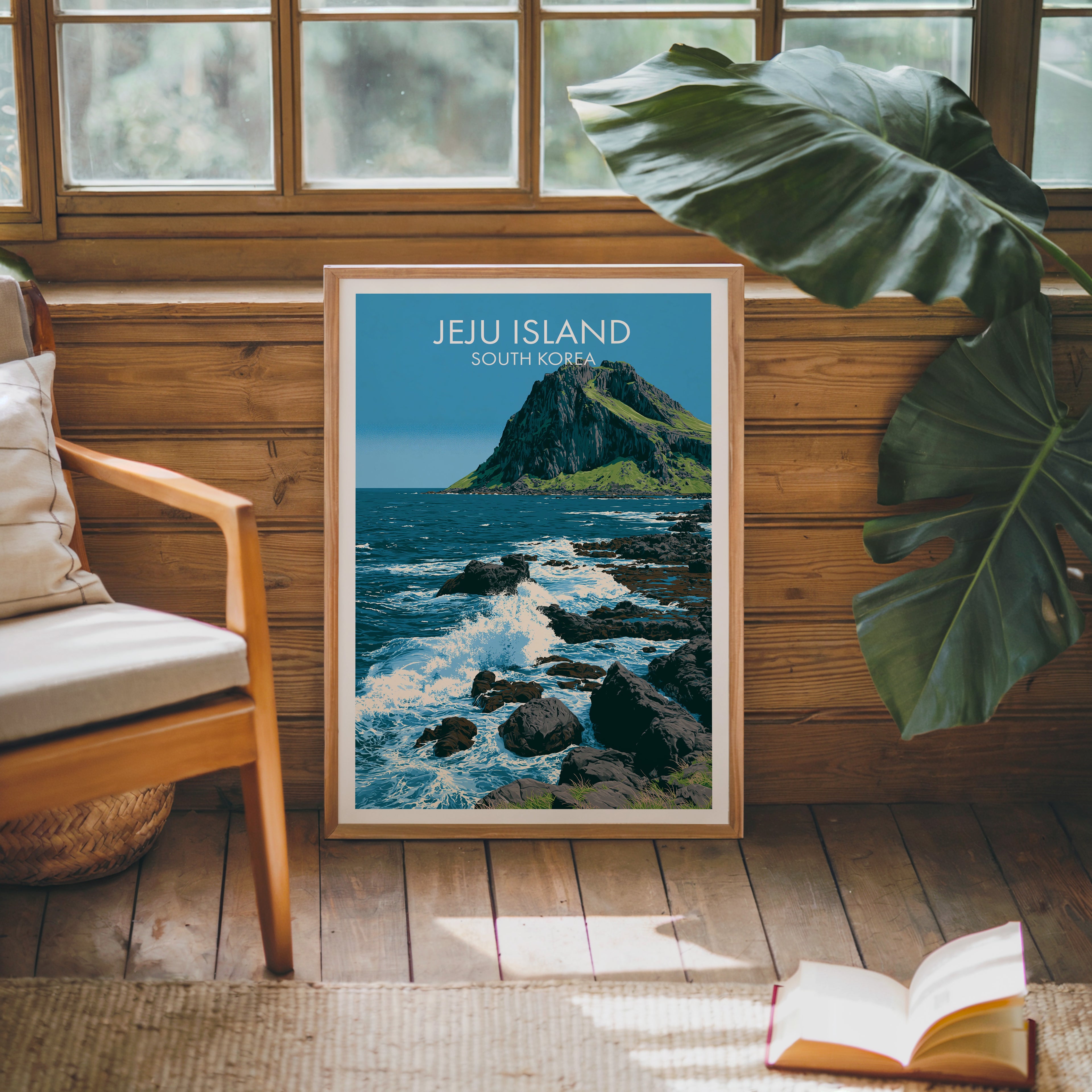Framed Seongsan Ilchulbong Jeju Korea poster leaning against a timber cabin wall with monstera leaves and an open book