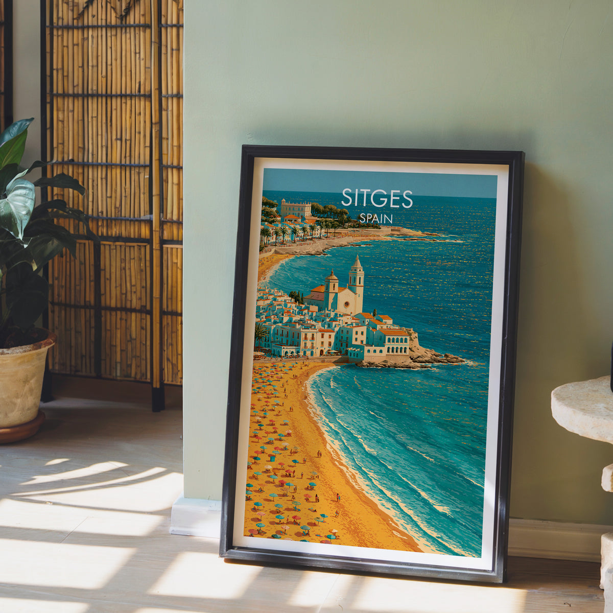 Sitges headland church poster, dark frame leaning on a sage-green wall beside a bamboo screen and a peace lily