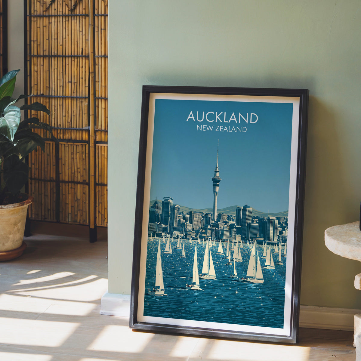 Auckland yacht harbour artwork in a black frame on a pale wood floor by a sage-green wall and bamboo screen