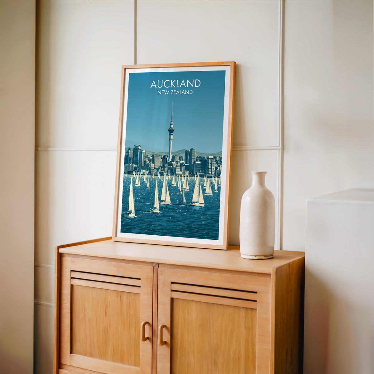 Auckland print in an oak frame on a light oak cabinet beside a pale bottle vase and cream panelled wall