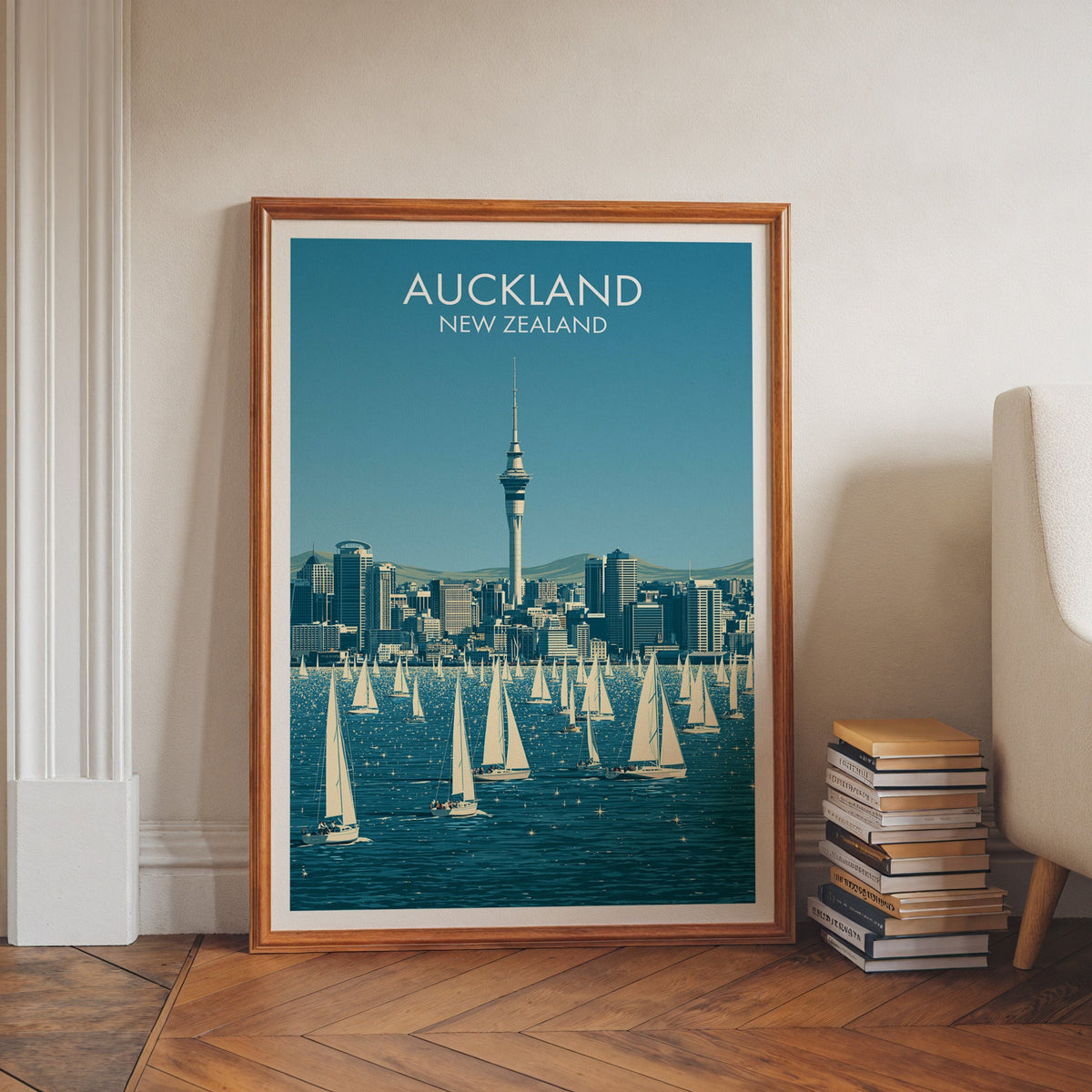 Auckland city print in a walnut frame leaning on herringbone floor beside stacked books and a pale armchair