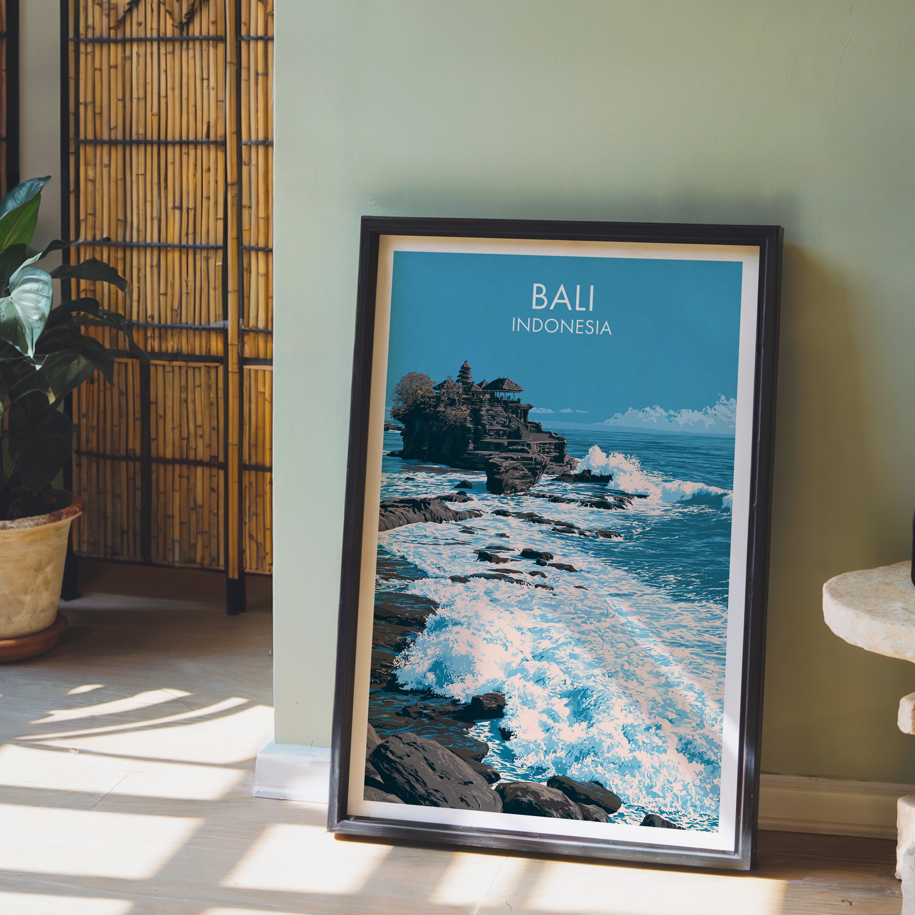 Tanah Lot Bali Indonesia wall art in a black frame leaning against a sage green wall beside a bamboo screen