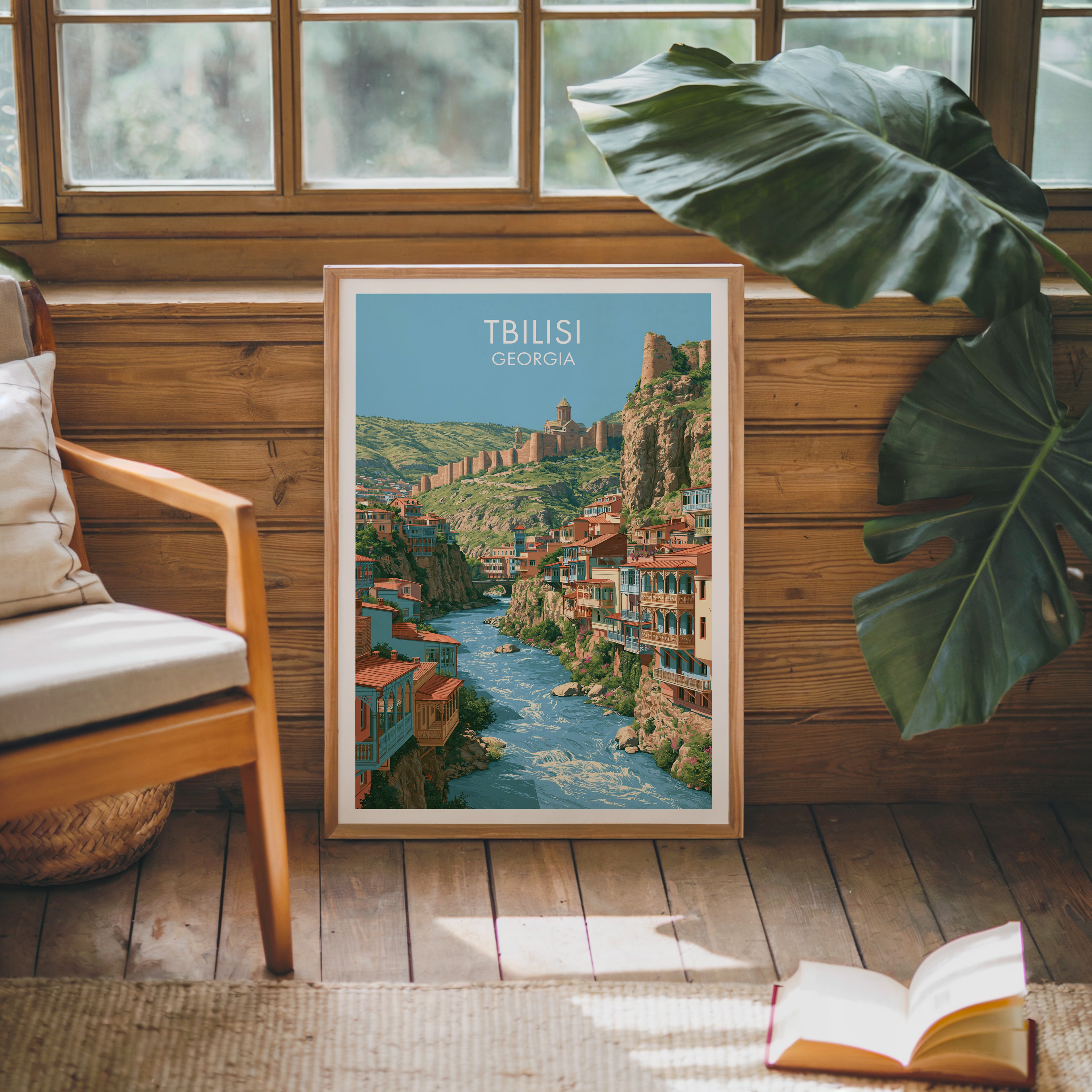 Framed Tbilisi Georgia poster leaning against a timber cabin wall with monstera leaves and an open book