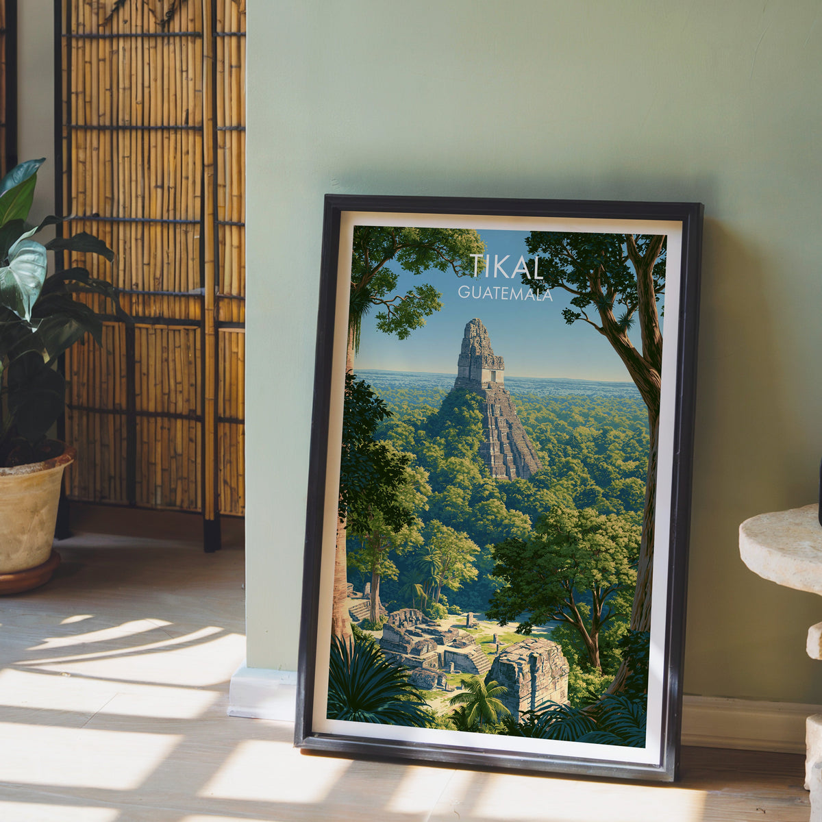 Layered green canopy print, black frame leaning on pale wood floor by a sage wall, bamboo screen and plant
