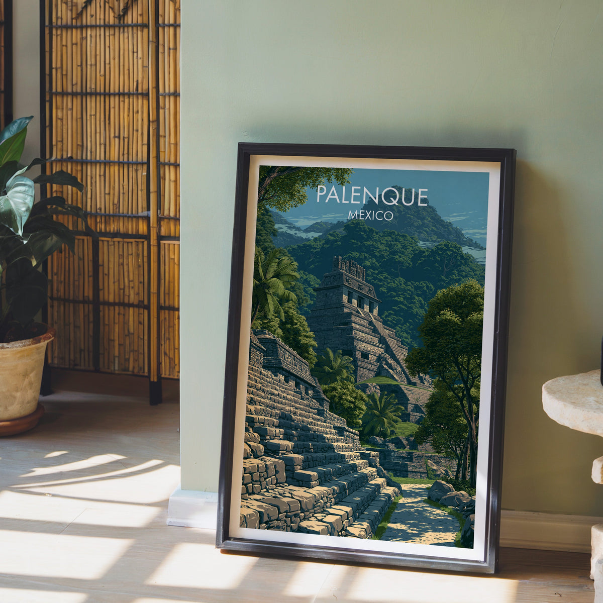 Green and blue-grey ruins print, black frame leaning on pale wood floor by a sage wall and bamboo screen