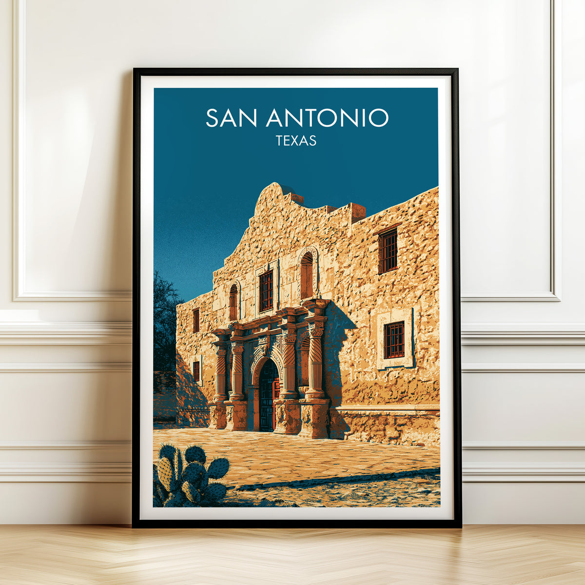 Alamo facade print in a black frame against panelled white wall trim above a herringbone wood floor.