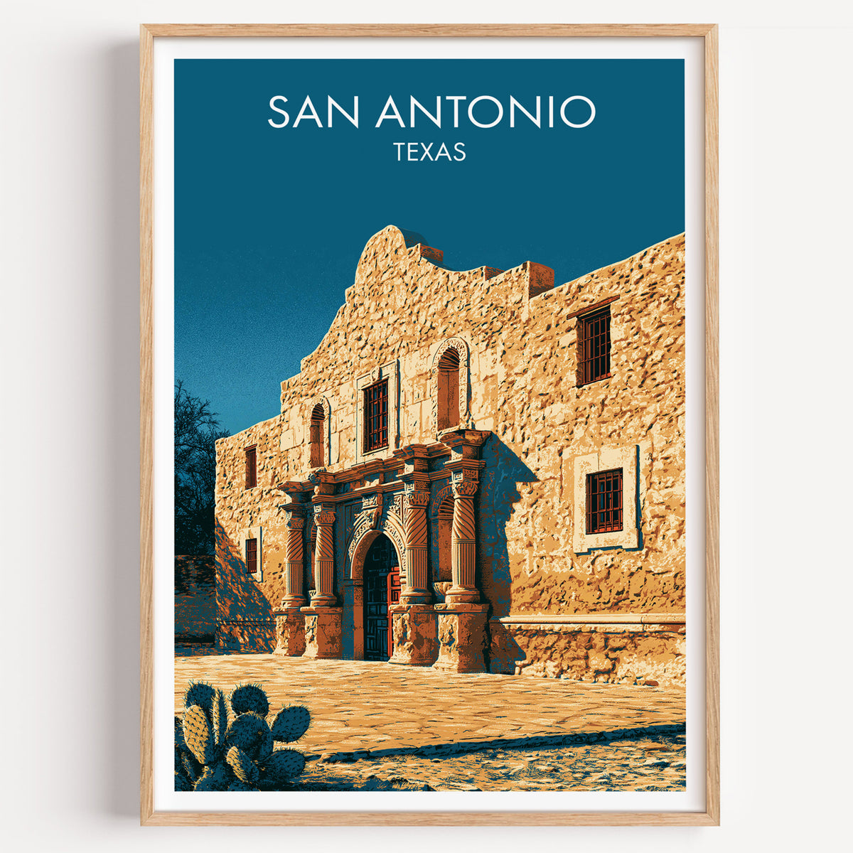 The Alamo travel poster in a light oak frame on a plain white wall, photographed straight on.