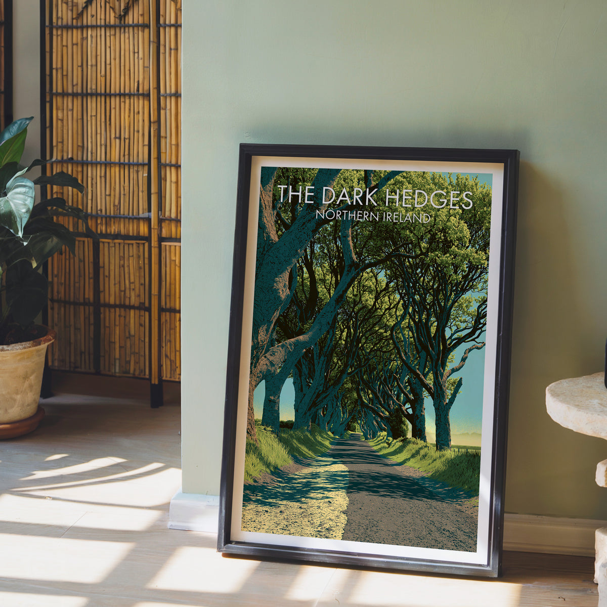 Dark Hedges Northern Ireland poster, dark frame leaning on a sage-green wall beside a bamboo screen and a peace lily