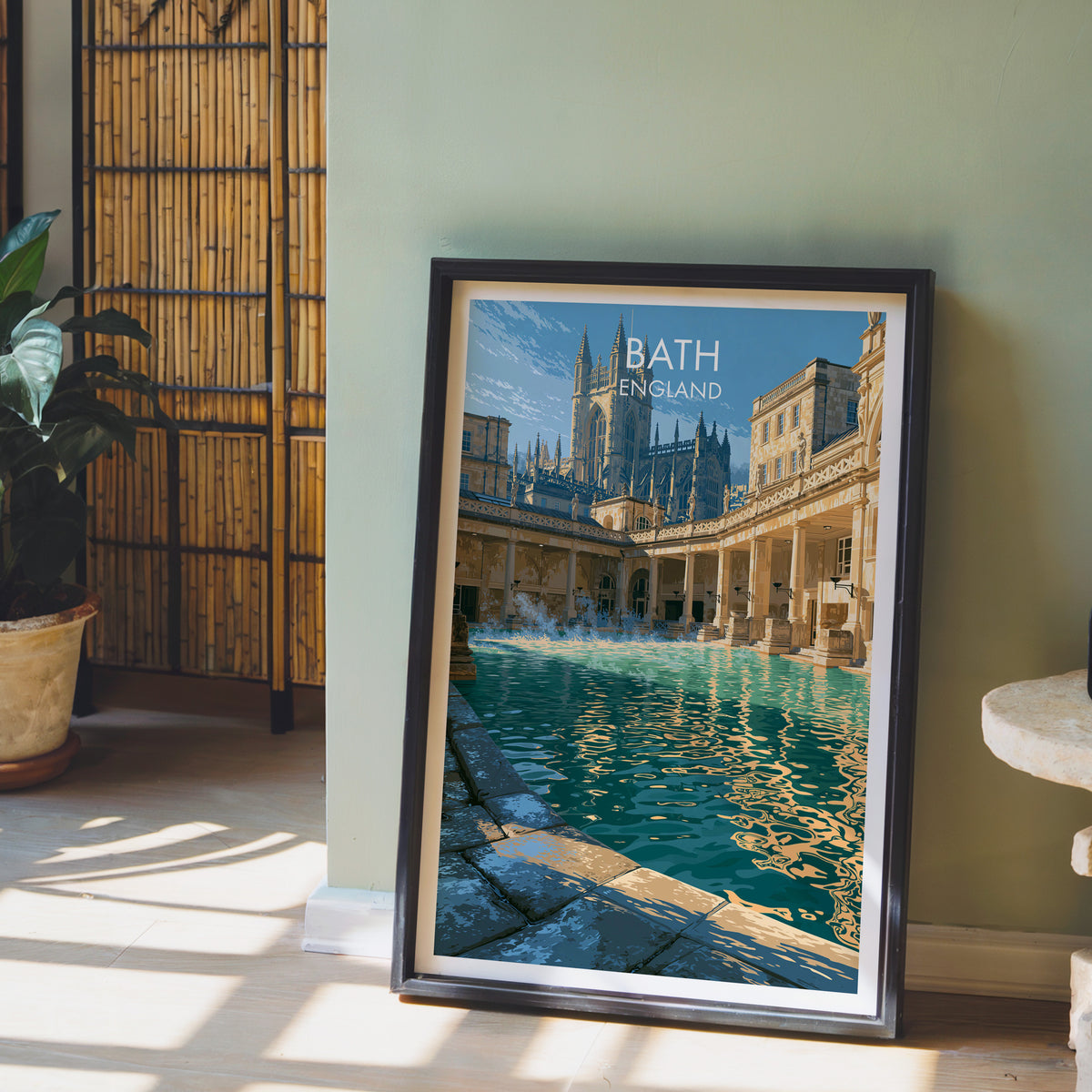 Bath Roman Baths poster, dark frame leaning on a sage-green wall beside a bamboo screen and a peace lily