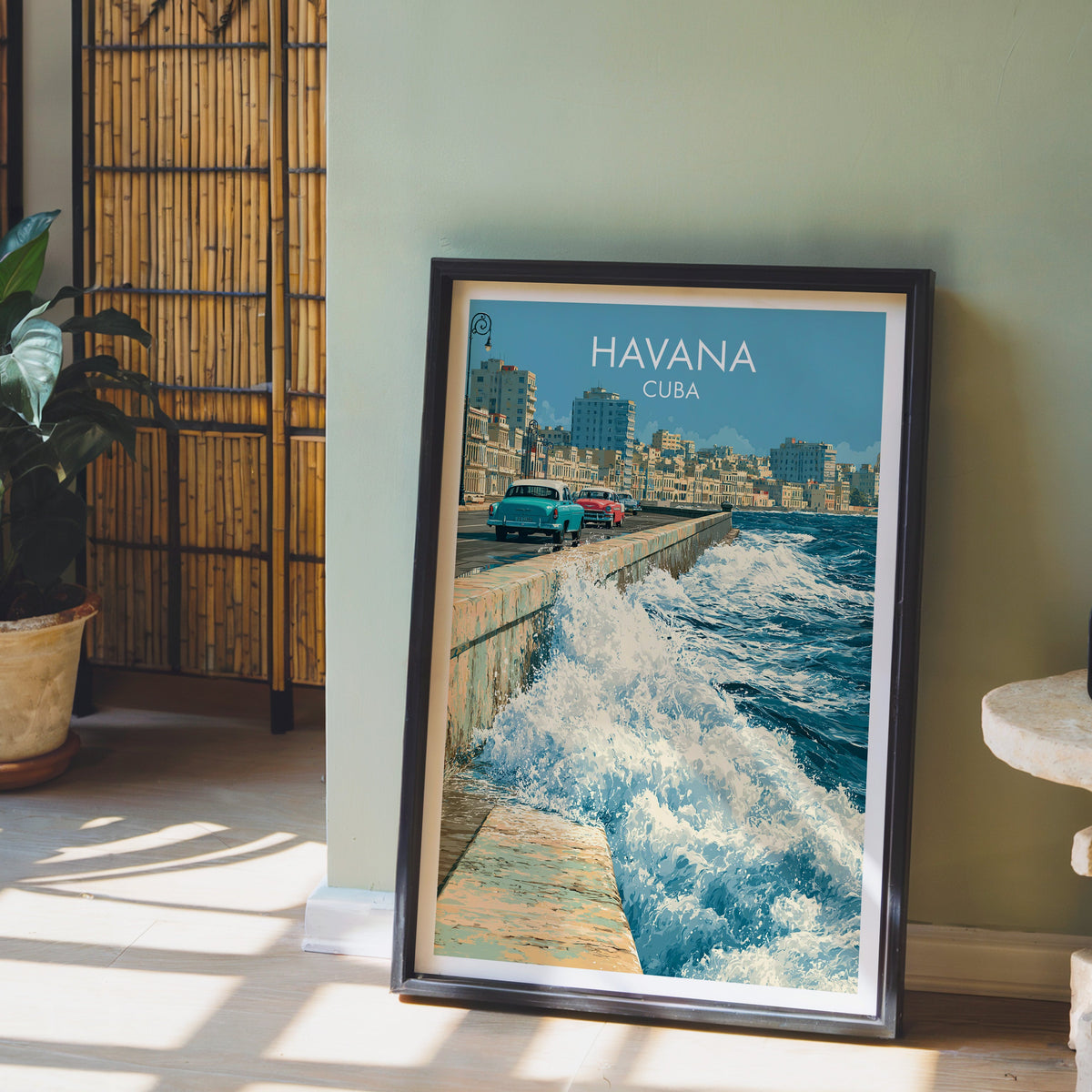 Blue ocean and seawall print in a black frame leaning on pale wood floor, sage-green wall, bamboo screen and plant