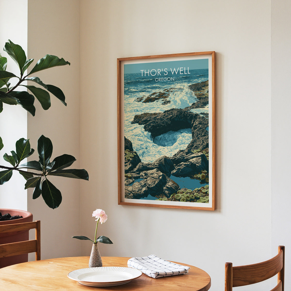 Walnut-framed Oregon coast poster in a dining corner with a round table, checked napkin and fiddle-leaf fig