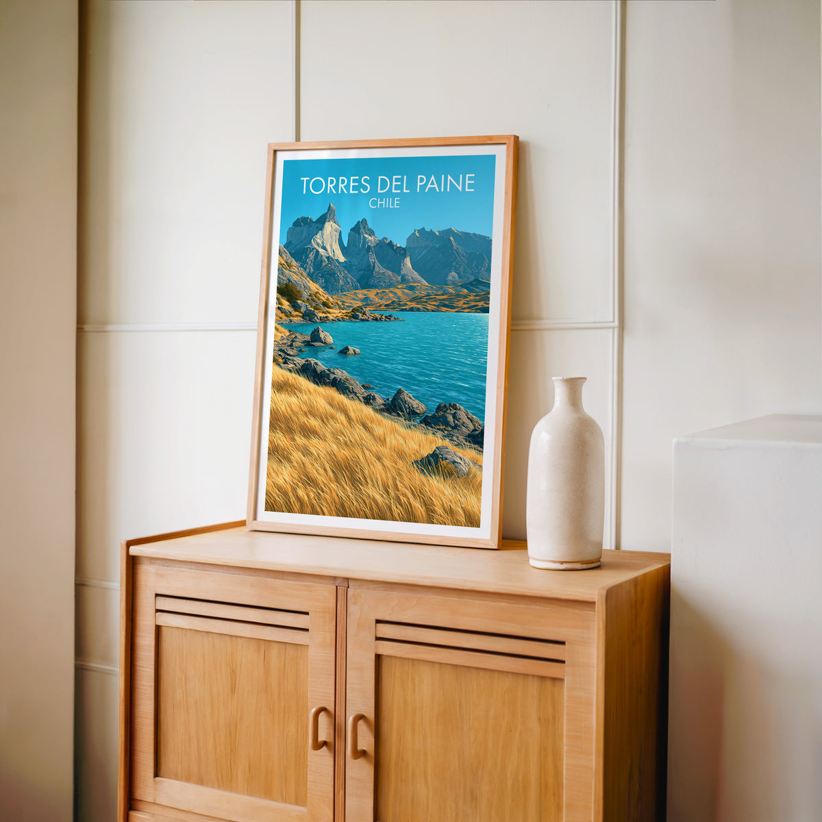 Golden grass and lake print in an oak frame on a pale oak cabinet against a cream panelled wall with a tall vase