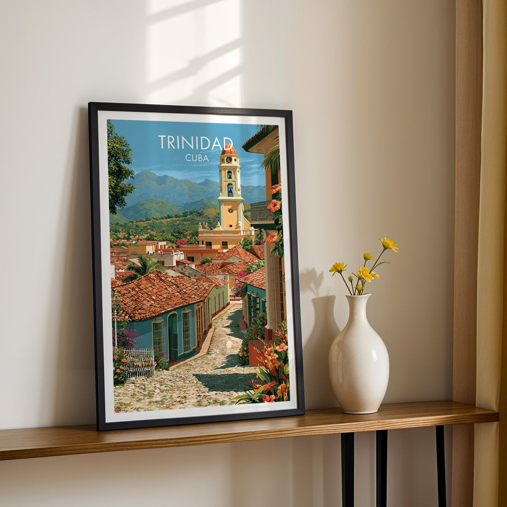 Terracotta rooftops of Trinidad in a black frame on a narrow wood shelf with yellow daisies and a sunlit cream wall