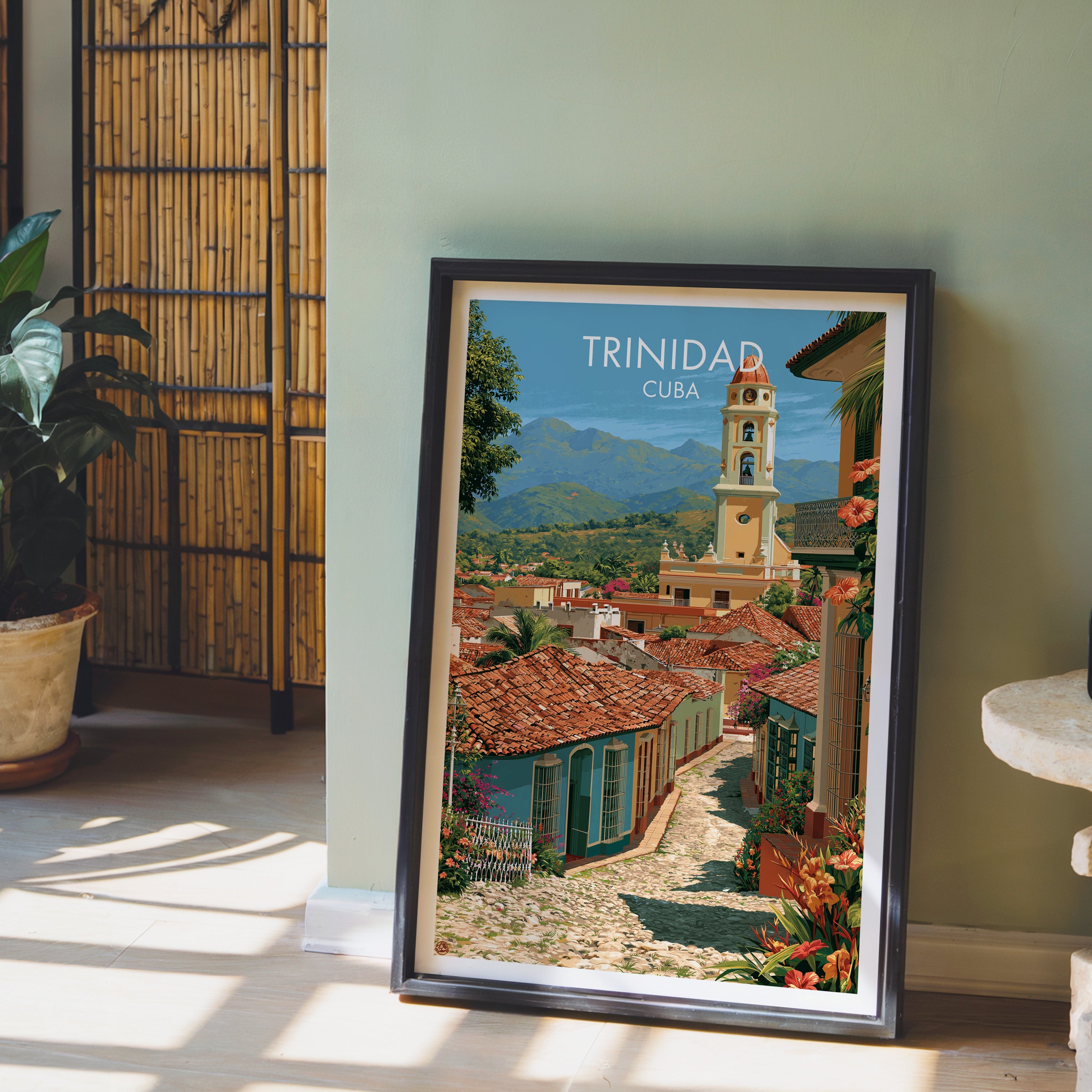 Cobbled street print in a black frame leaning on pale wood floor by a sage-green wall with a bamboo screen and plant