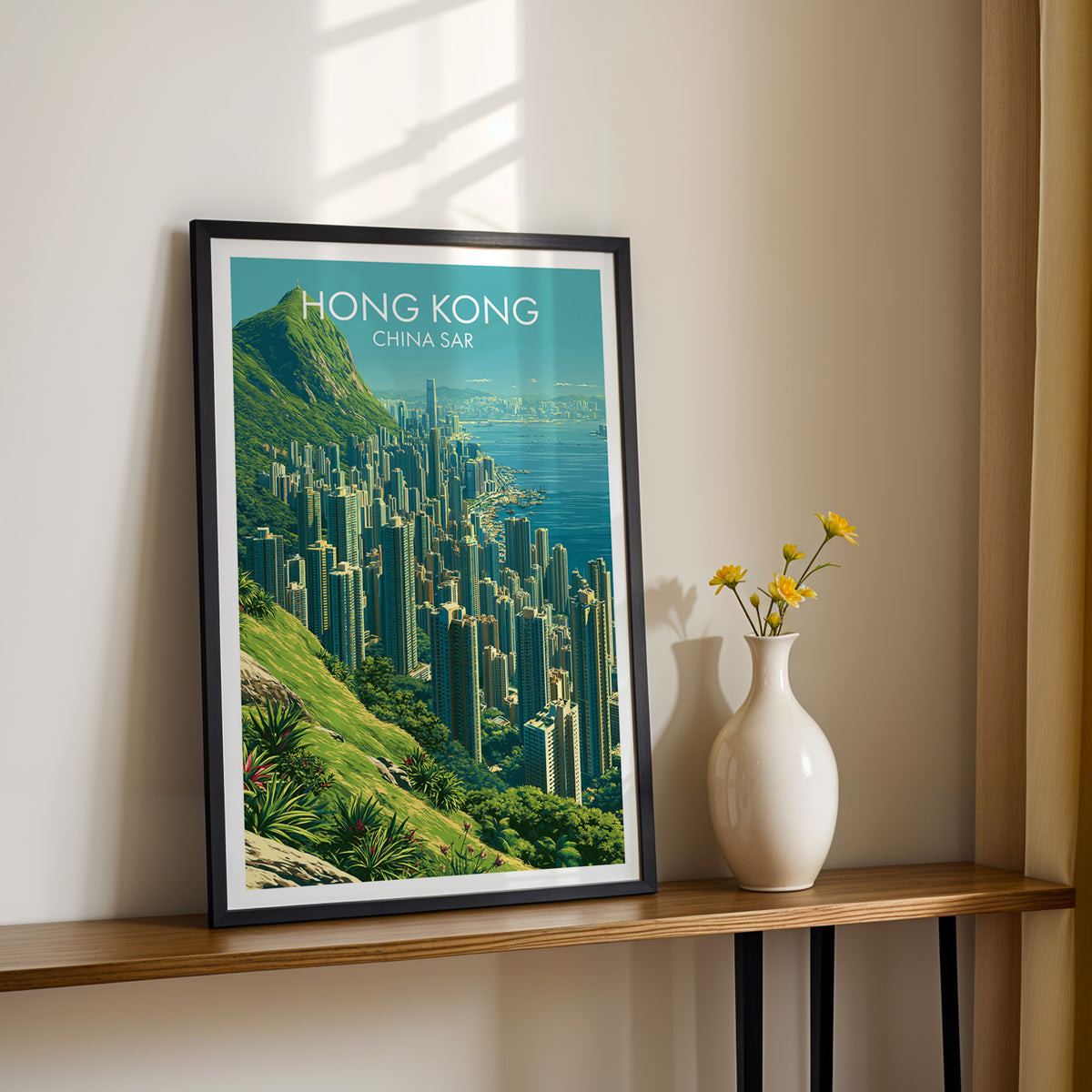 Victoria Peak hillside wall art in a black frame on a narrow wood shelf with yellow daisies, cream wall in sunlight