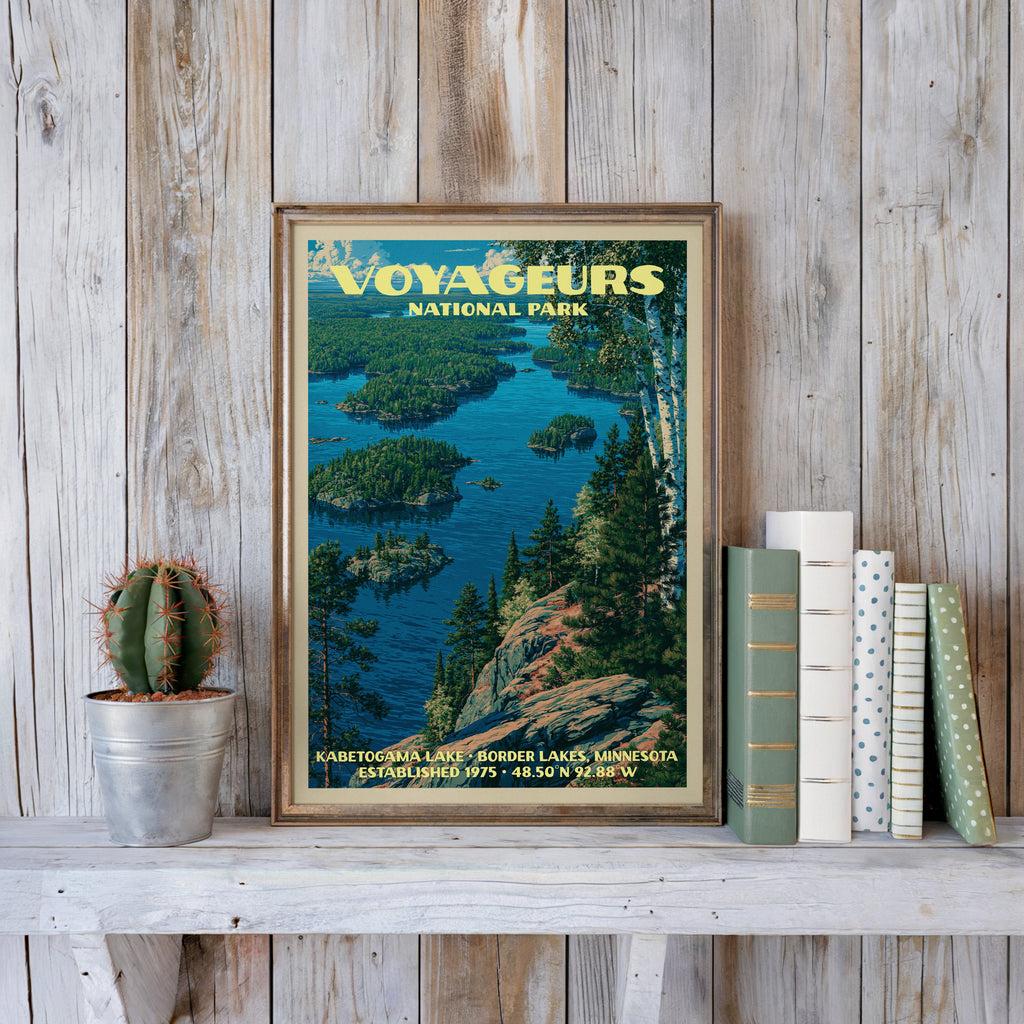 Voyageurs lake print in a slim gold frame on a whitewashed plank wall beside a cactus and pastel books