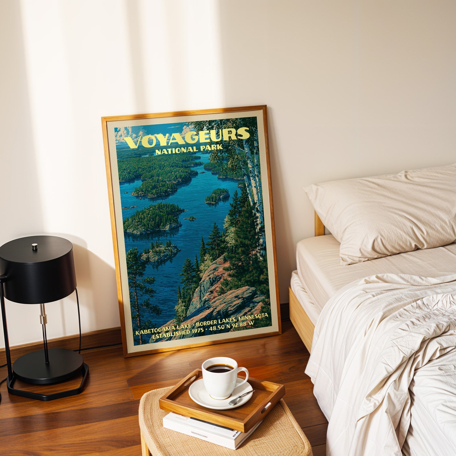 Kabetogama Lake Voyageurs print in a gold frame on the floor beside a made bed and a black table lamp