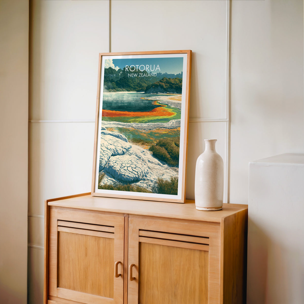 Rotorua print in an oak frame on a light oak cabinet beside a pale bottle vase and panelled wall