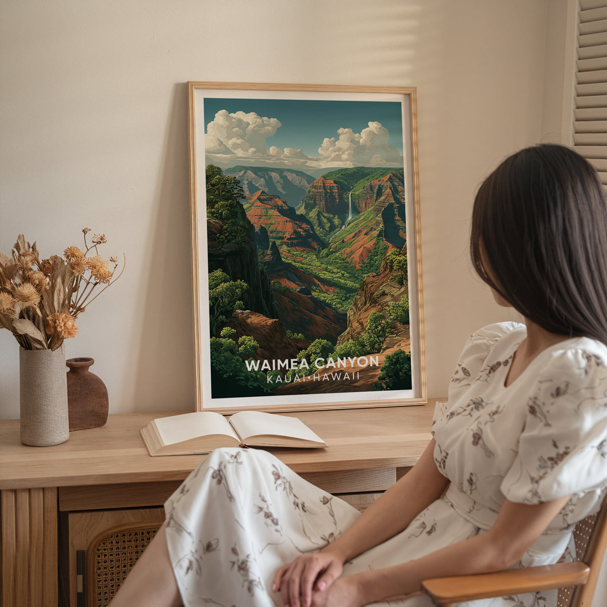 Waimea Canyon print on a wood sideboard with dried flowers and an open book, a woman seated in white