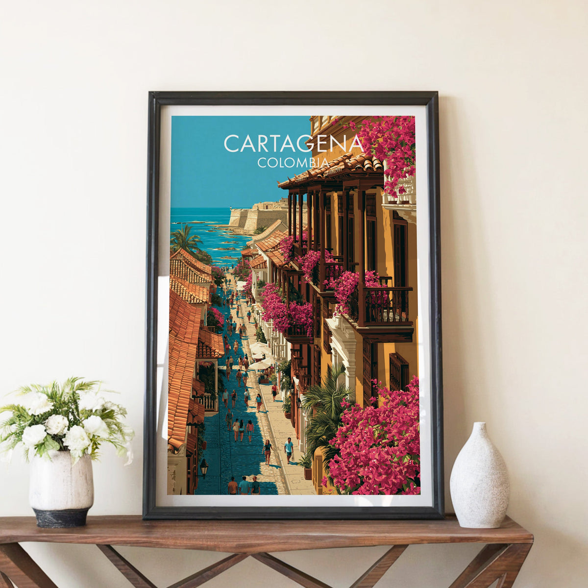 Walled city street poster in a black frame on a brown X-leg console with white blooms and a ceramic vase