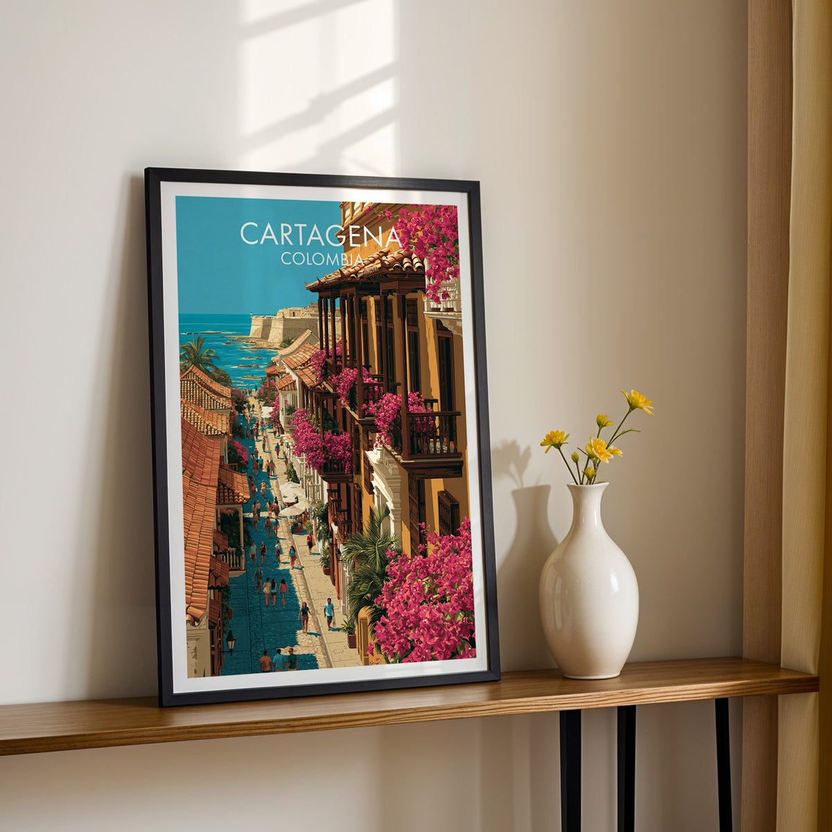 Cartagena balcony print in a black frame on a slim wood shelf with yellow daisies in a white vase, cream wall