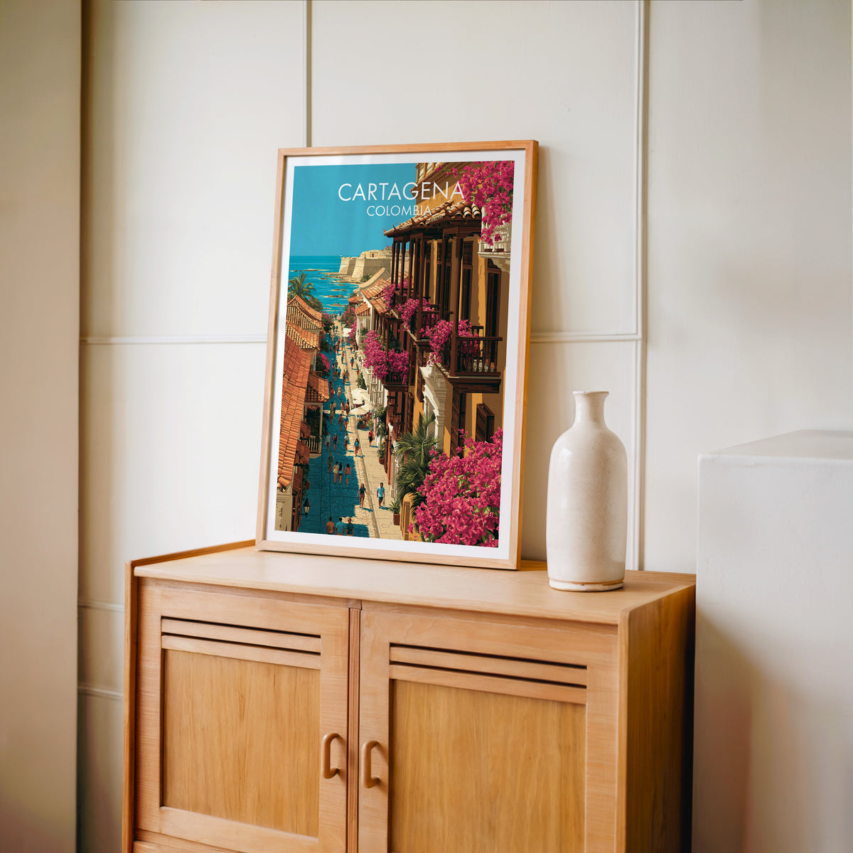 Cartagena travel poster in an oak frame on a light oak cabinet against a cream panelled wall with a tall vase