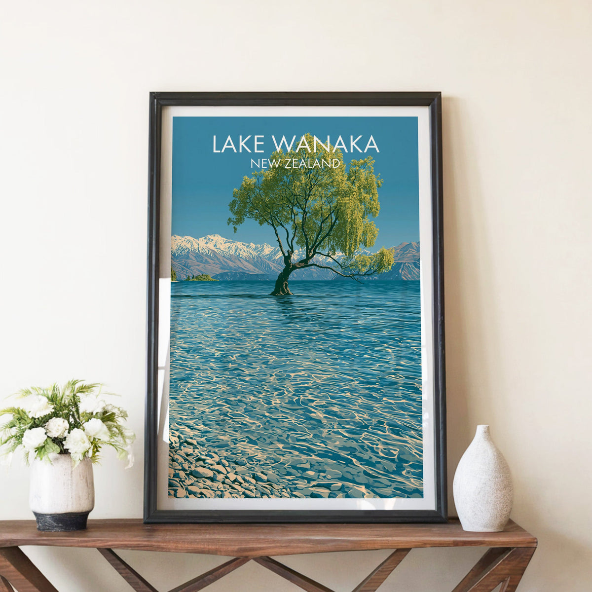 Lake Wanaka poster in a black frame on a walnut console with white peonies and a tall ceramic vase