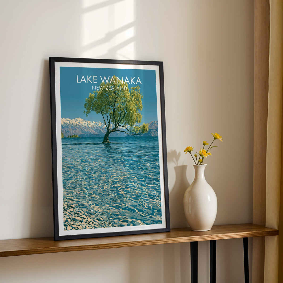 Lone tree in blue water print, black frame on a wood shelf with yellow daisies on a sunlit cream wall