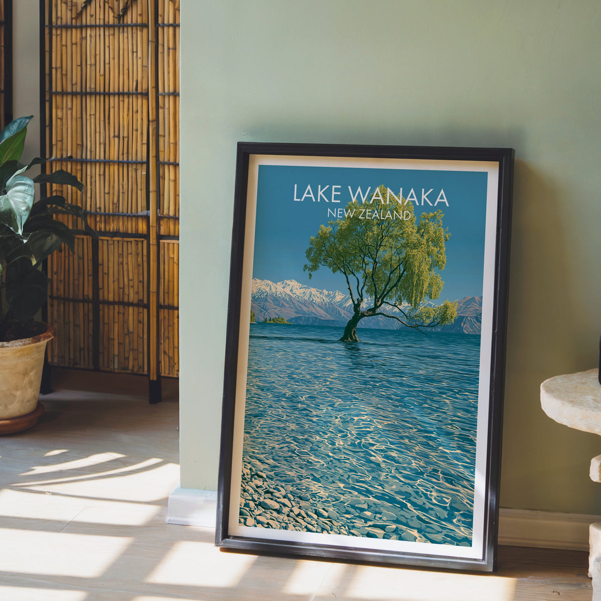 New Zealand lake and peaks artwork in a black frame on a pale wood floor by a sage wall and bamboo screen