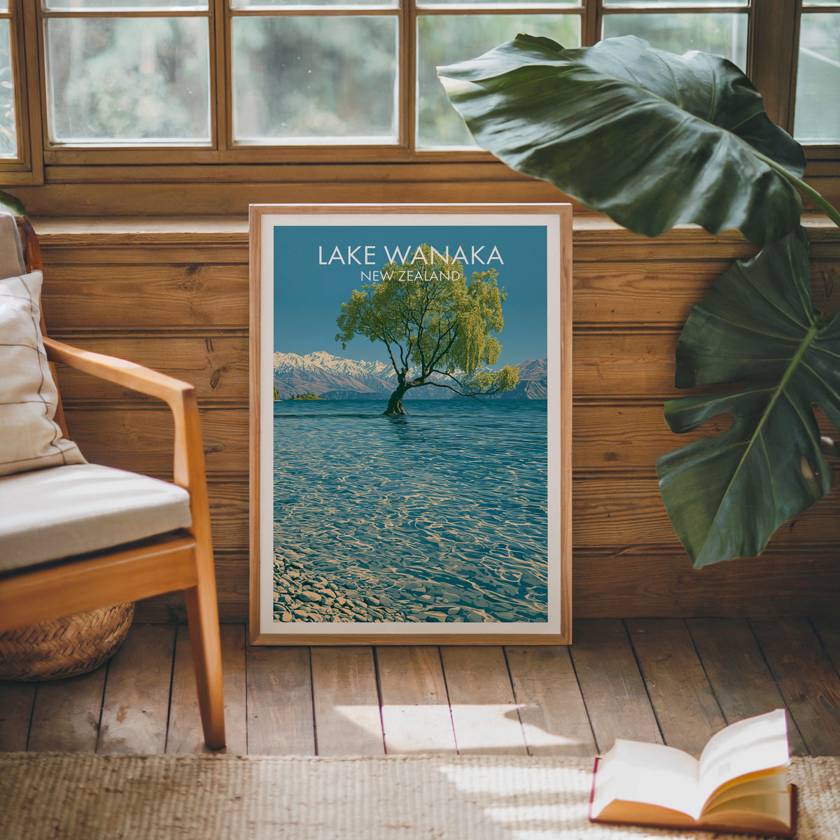 Lone willow print leaning on a pine cabin wall beside a wooden armchair, monstera leaf and open book