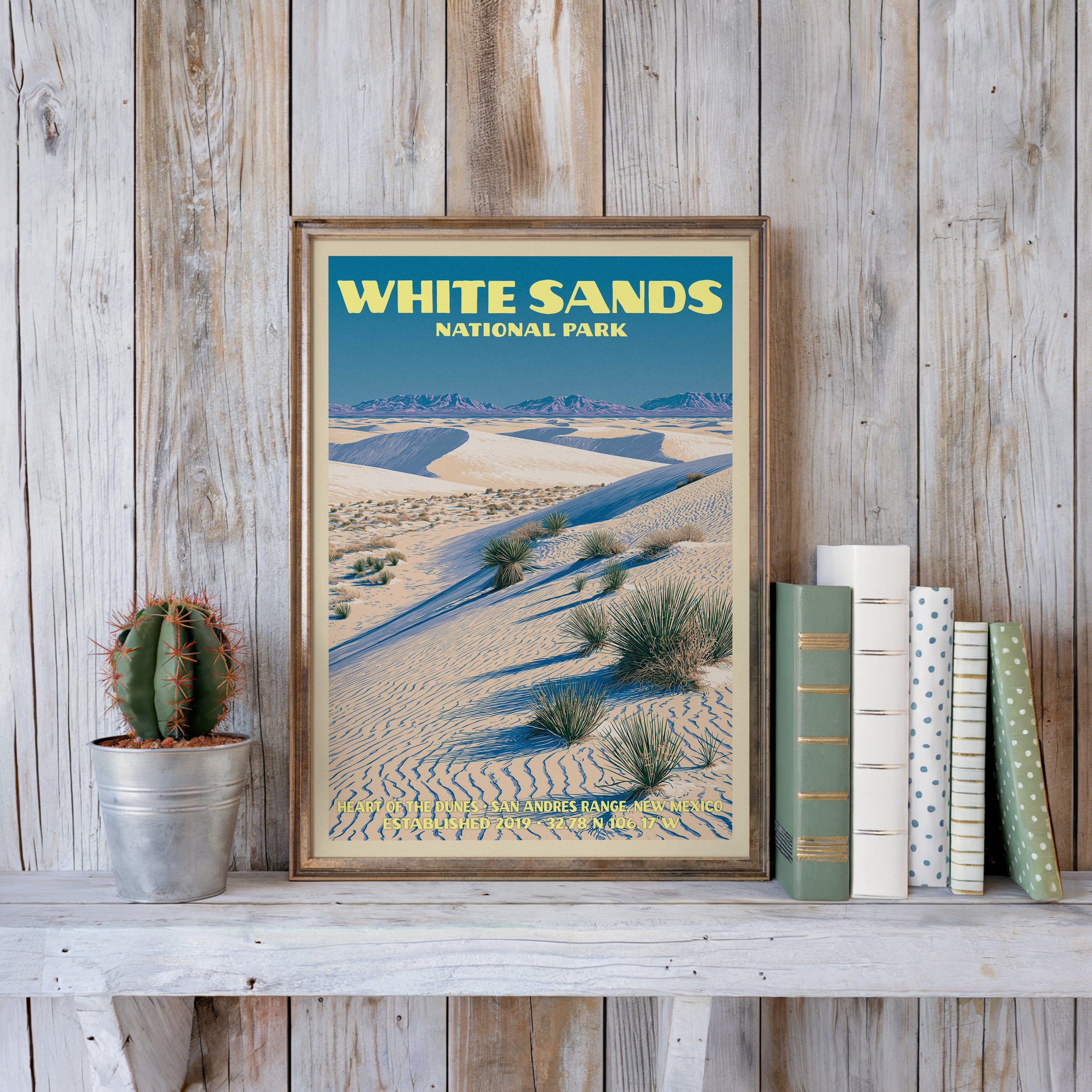 White Sands vintage poster in a slim gold frame on a whitewashed plank wall beside a cactus and pastel books