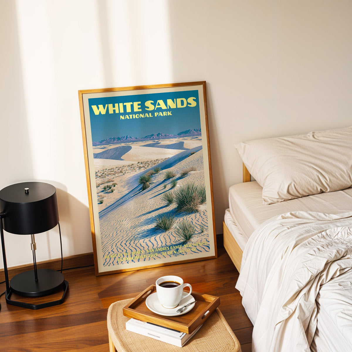Heart of the Dunes poster in a gold frame on the bedroom floor beside a made bed and a black table lamp