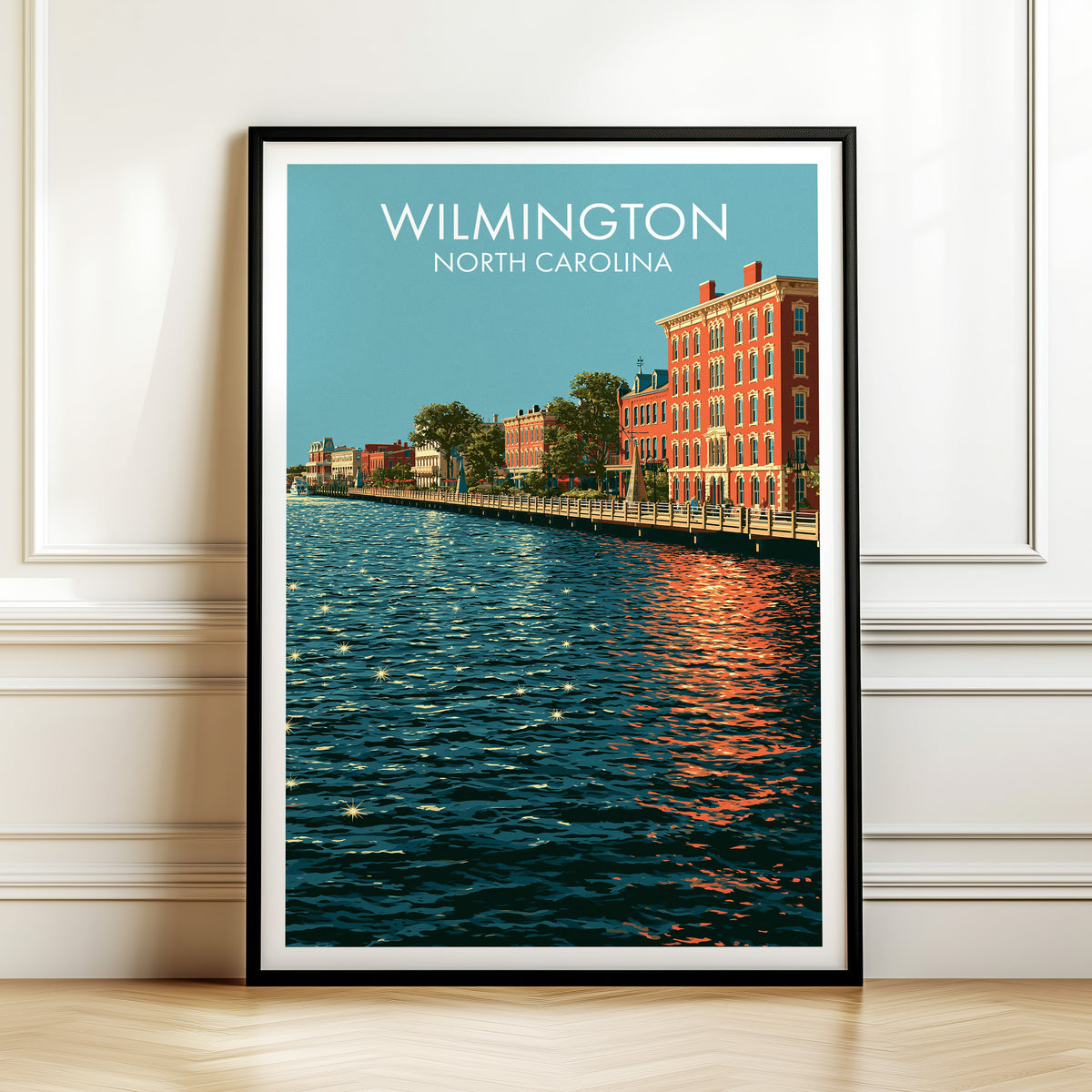 Wilmington Riverwalk print in a black frame leaning on white panelled moulding above a herringbone floor.