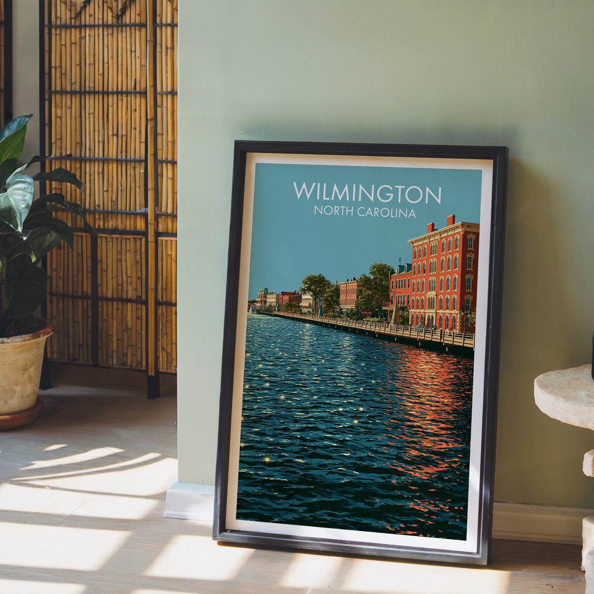 Riverfront boardwalk poster in a black frame on a sage green wall with bamboo screen and a potted peace lily.