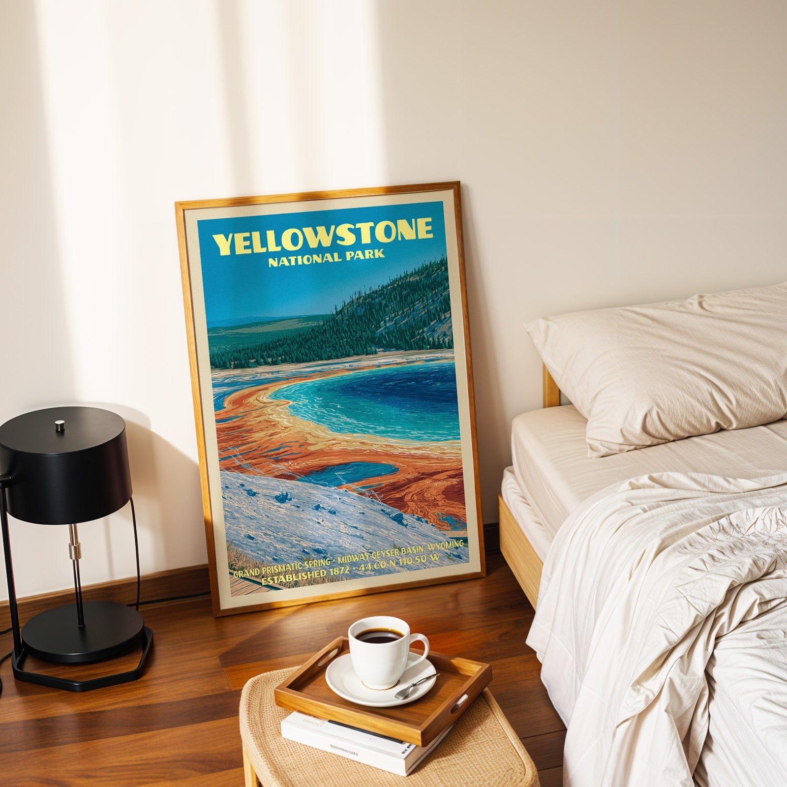 Grand Prismatic Spring poster in a gold frame on the bedroom floor beside a made bed and a black lamp
