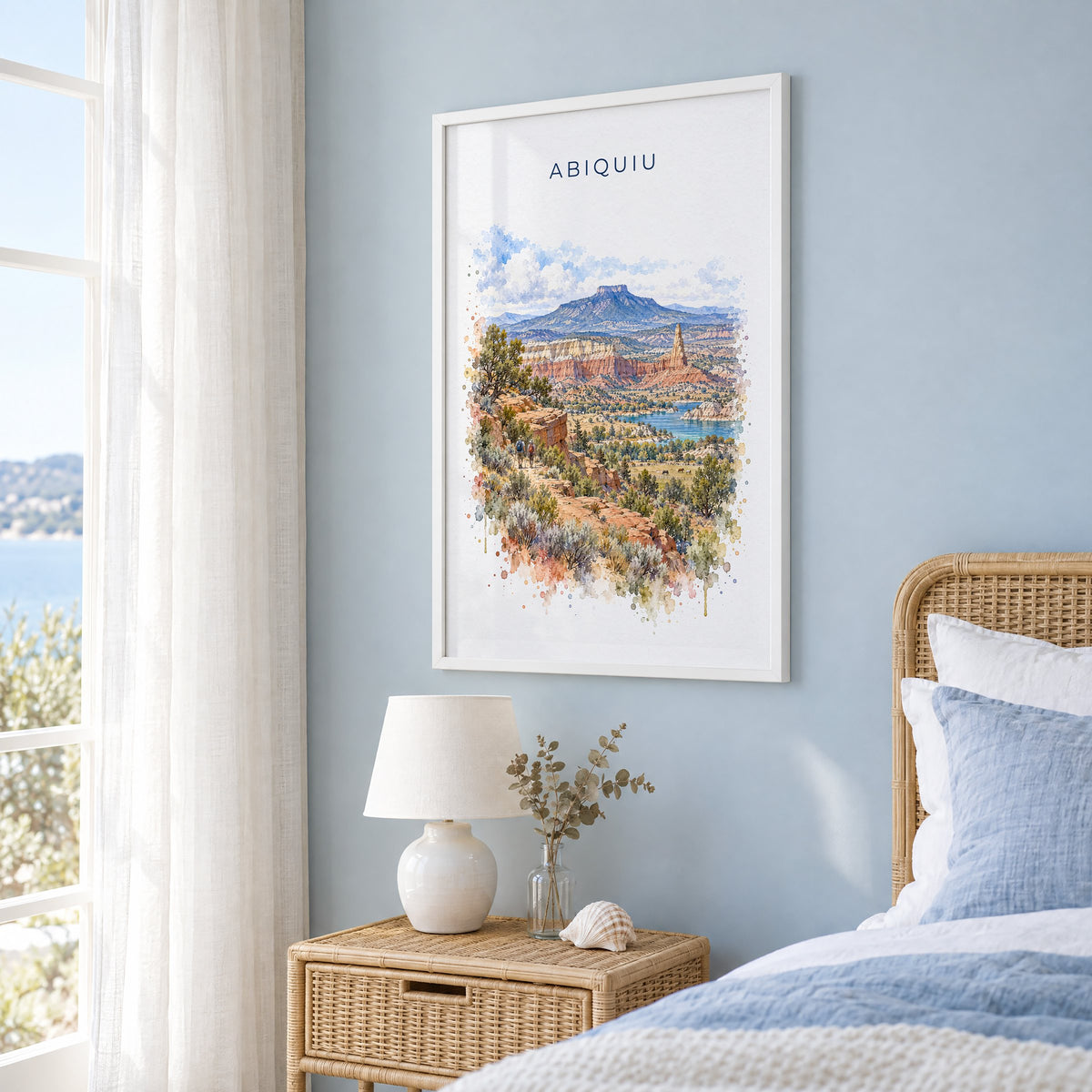 Abiquiu, New Mexico watercolor print in a white frame on a pale blue wall beside a rattan bed in a coastal bedroom