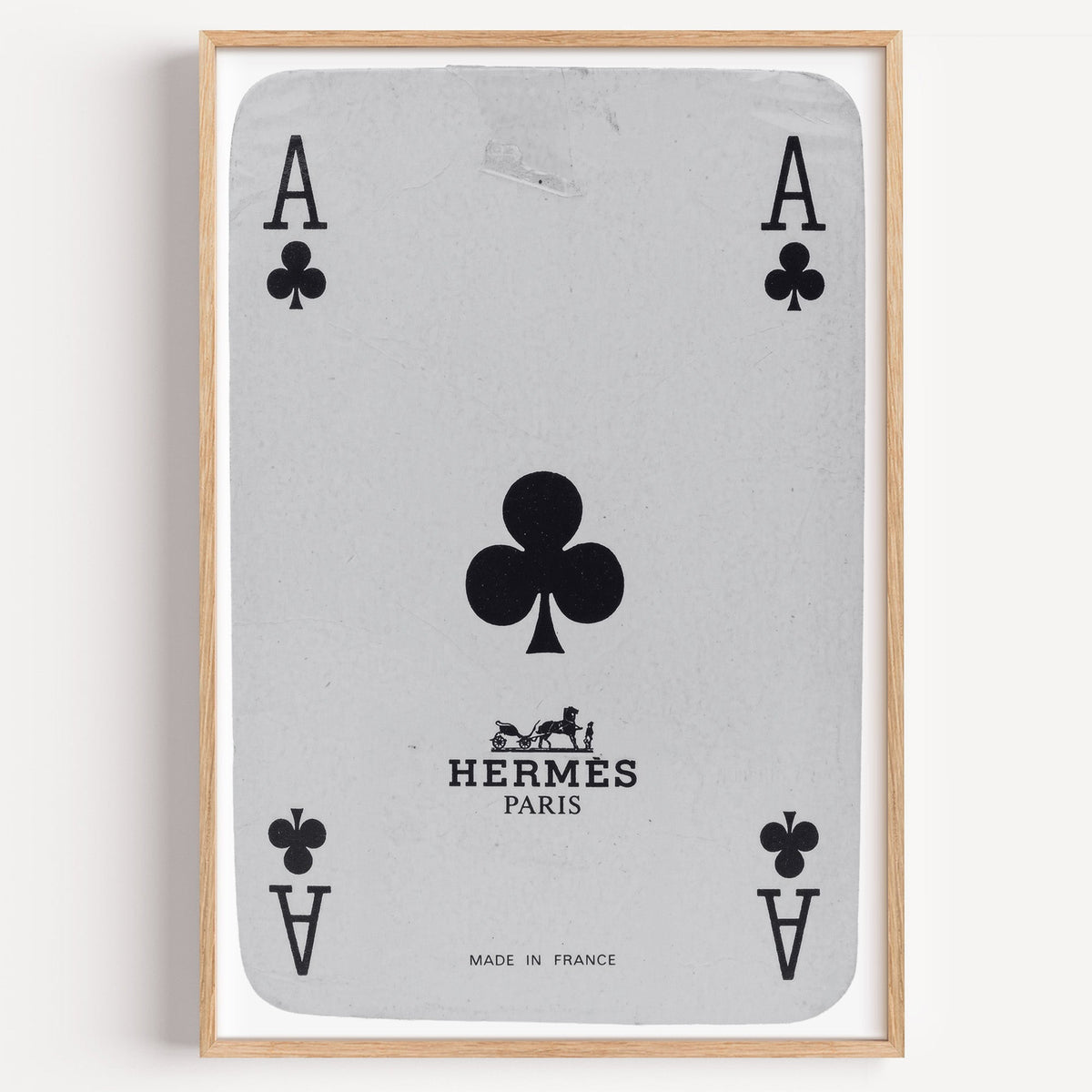 Worn vintage ace of clubs playing card print in a light oak frame on a plain white wall