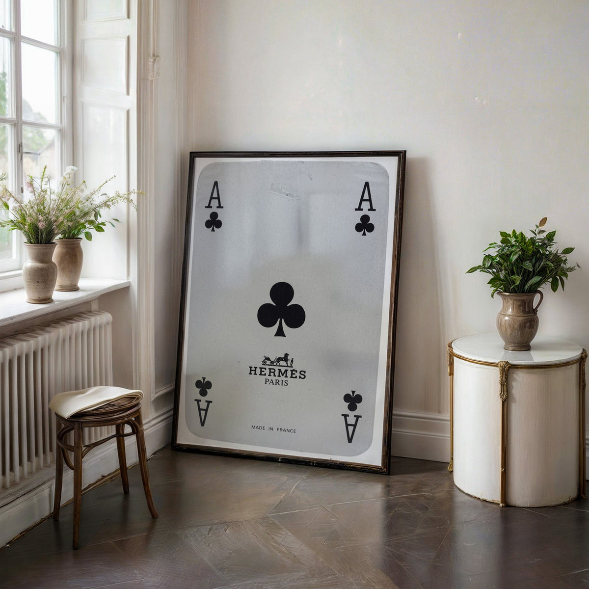 Worn vintage ace of clubs playing card print in a black frame on a dark stone floor beside a white side table