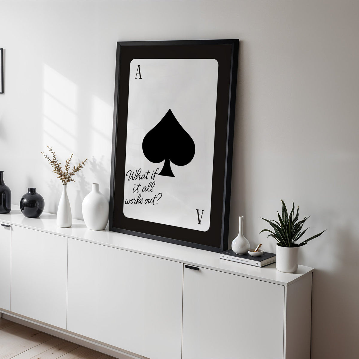 Ace of spades card with What if it all works out quote print in a black frame on a white sideboard with black and white vases