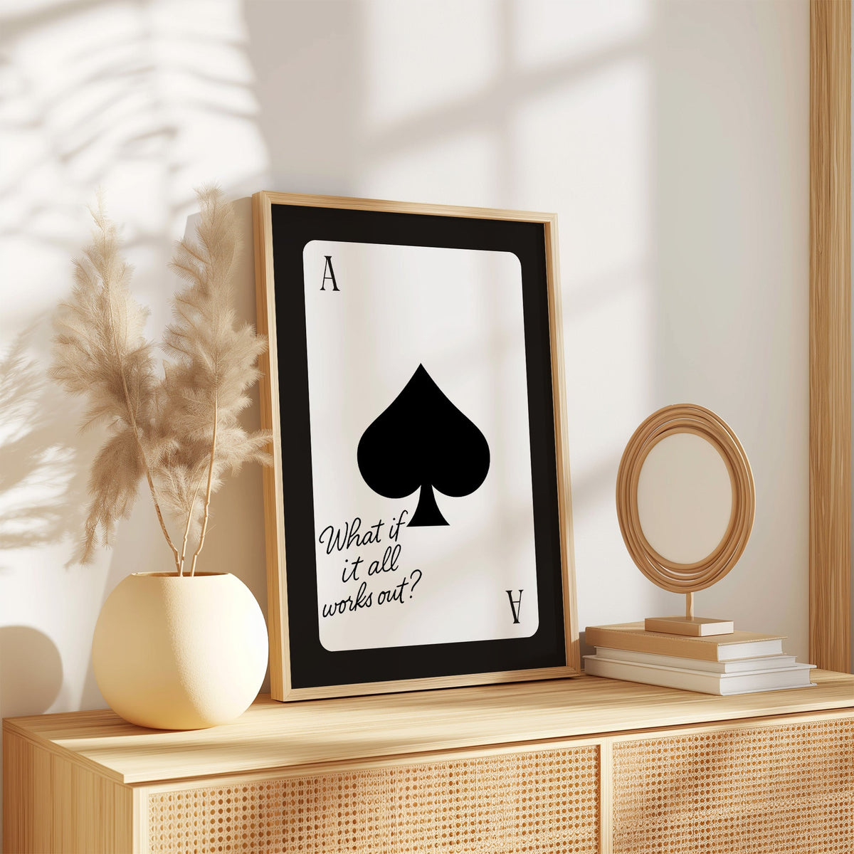 Ace of spades card with What if it all works out quote print in an oak frame on a rattan sideboard beside pampas grass