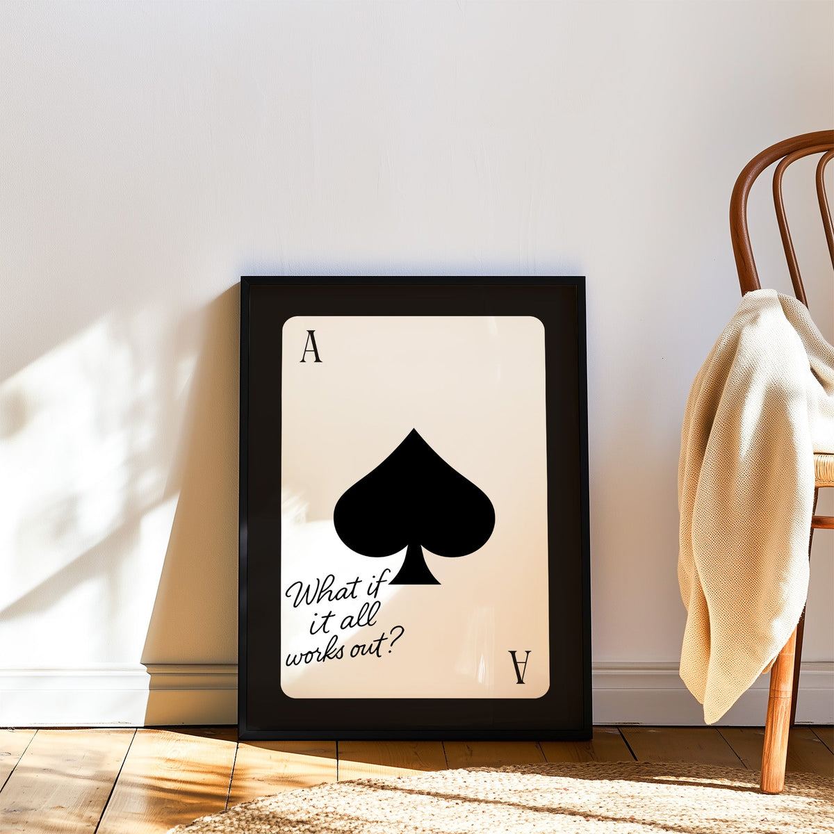 Ace of spades card with What if it all works out quote print in a black frame leaning on a wood floor beside a bentwood chair