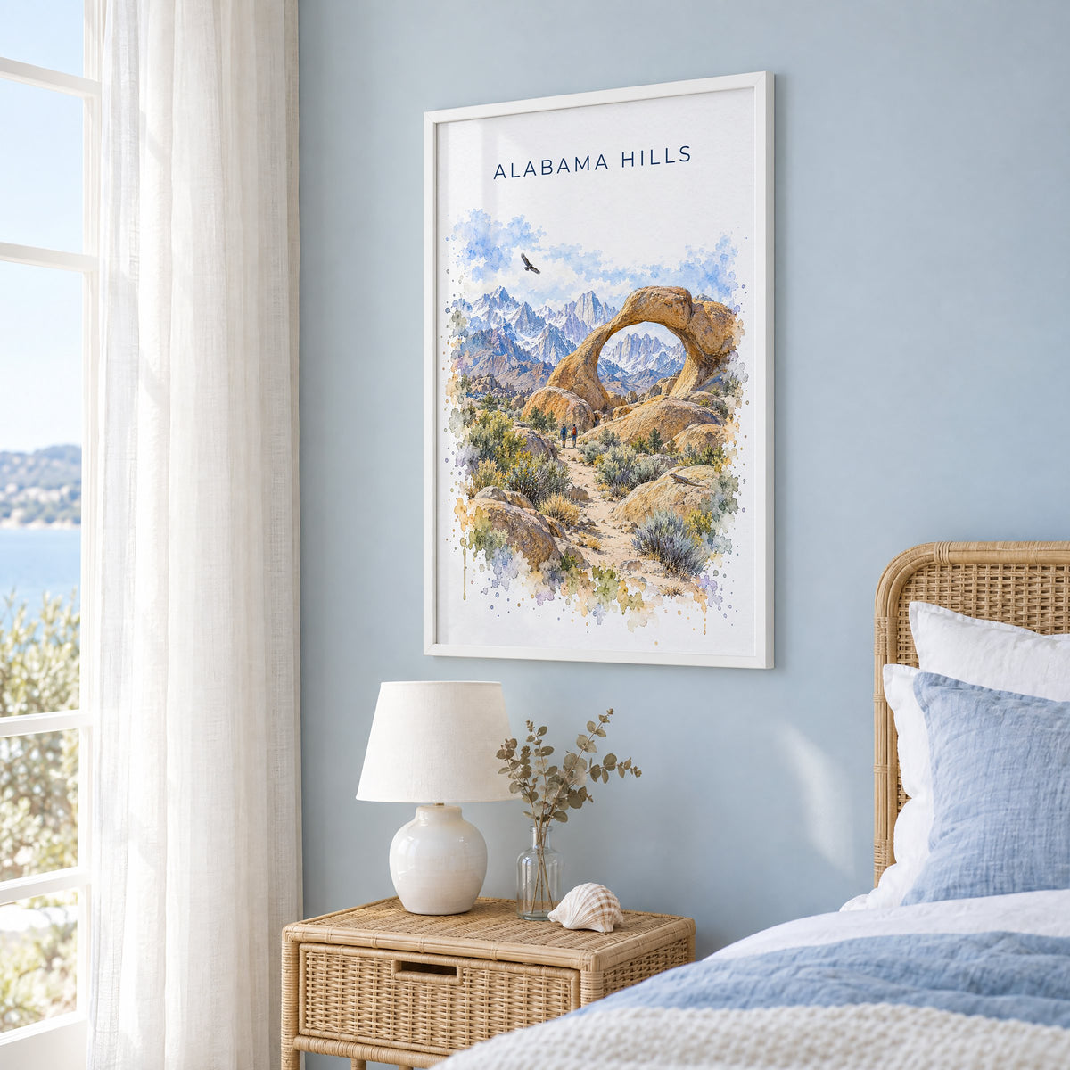 Alabama Hills watercolor print in a white frame on a pale blue wall beside a rattan bed in a coastal bedroom