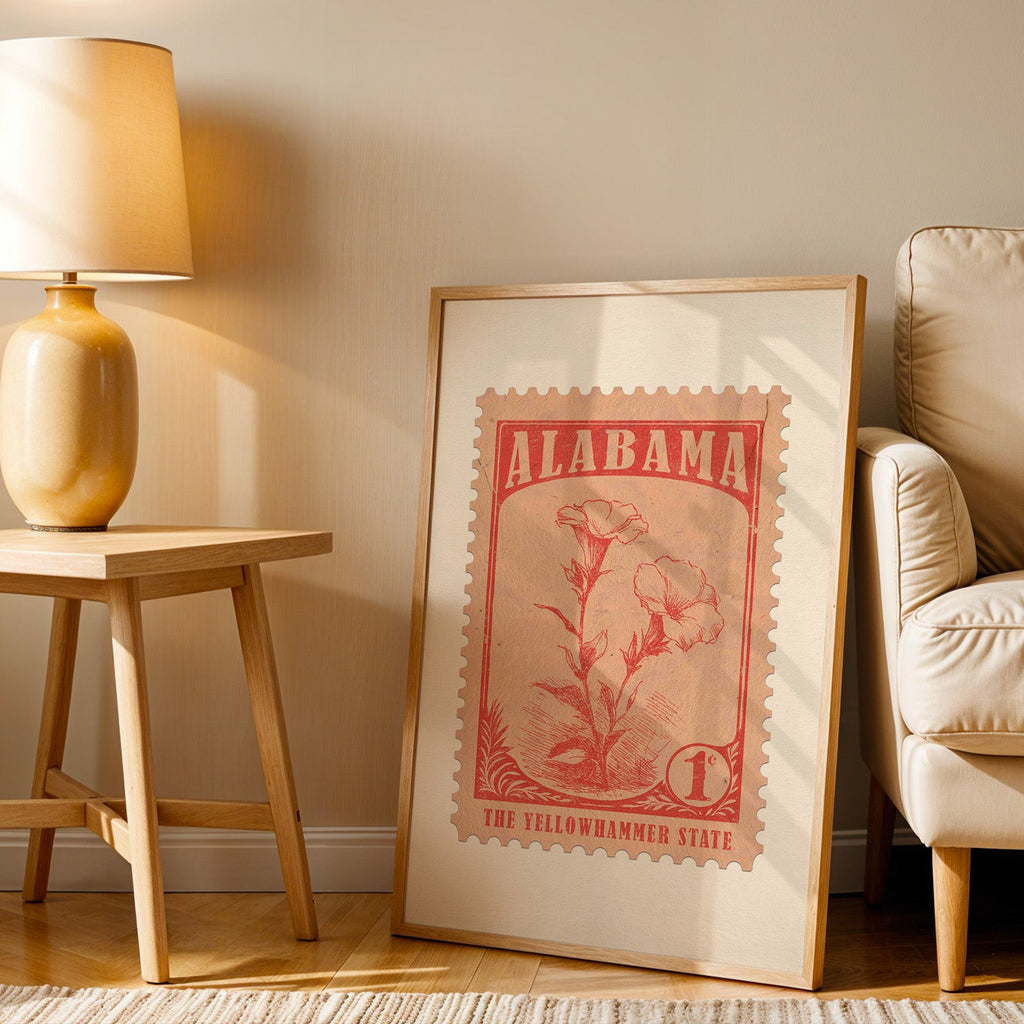 Alabama stamp print in a wood frame resting on the floor beside a beige armchair and a yellow table lamp