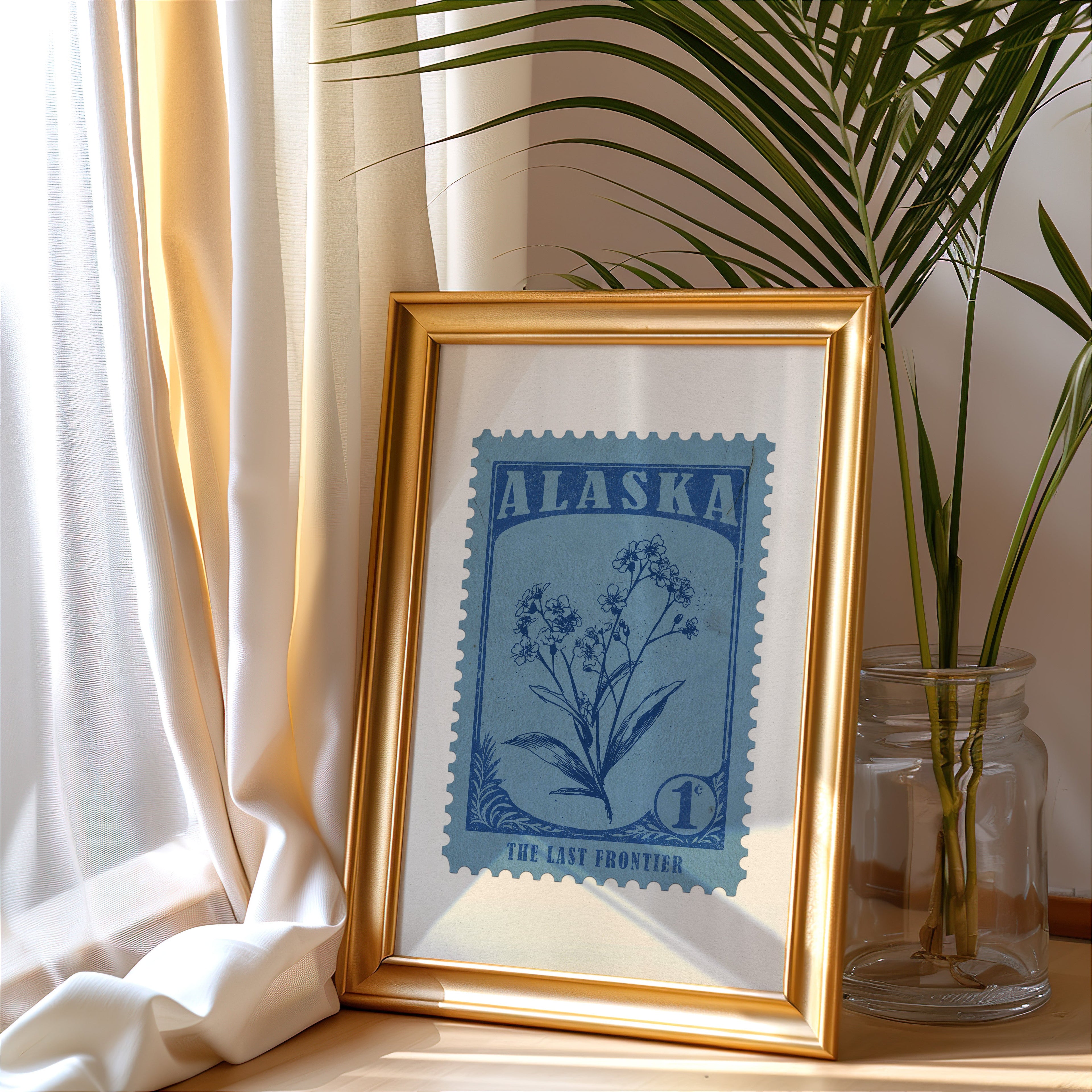 Alaska stamp print in a gold frame leaning by a cream curtain with a palm in a glass jar
