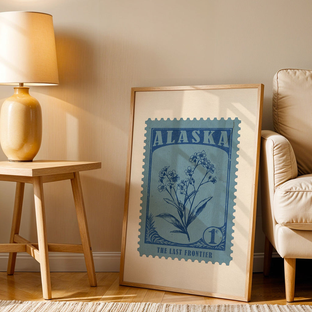 Alaska stamp print in a wood frame resting on the floor beside a beige armchair and a yellow table lamp