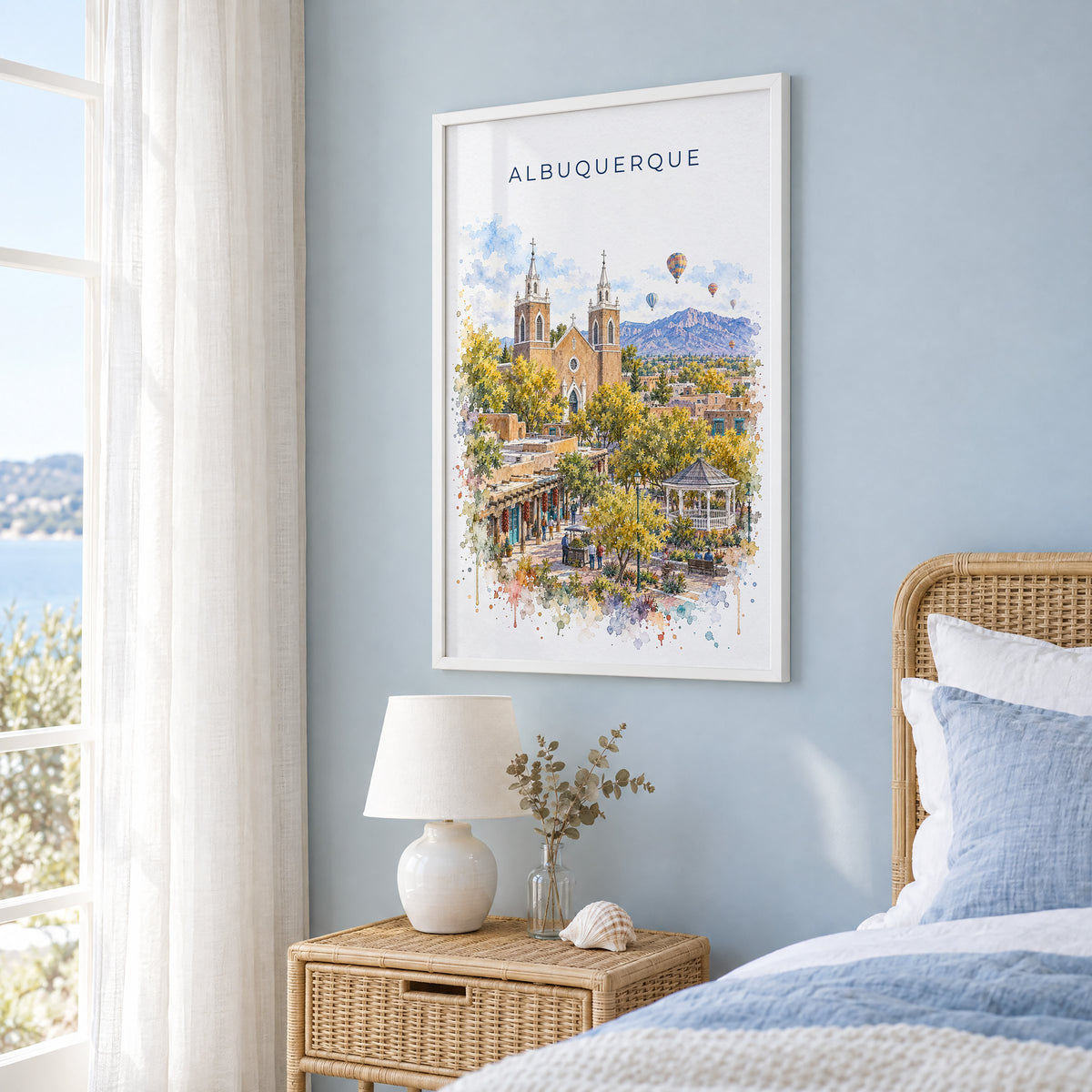 Albuquerque watercolor print in a white frame on a pale blue wall beside a rattan bed in a coastal bedroom